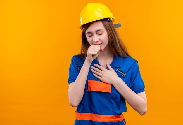 Young woman builder worker in construction uniform and safety helmet looking unwell coughing standing