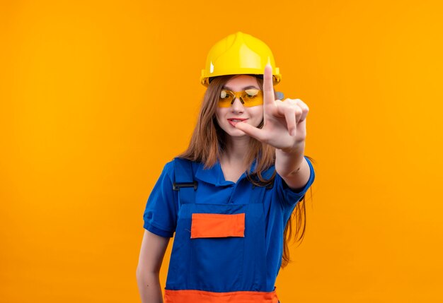 Young woman builder worker in construction uniform and safety helmet smiling showing index finger at front