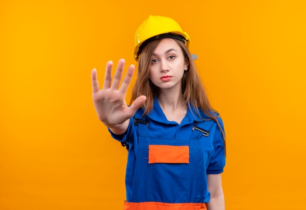 Young woman builder worker in construction uniform and safety helmet standing with open hand making stop sign