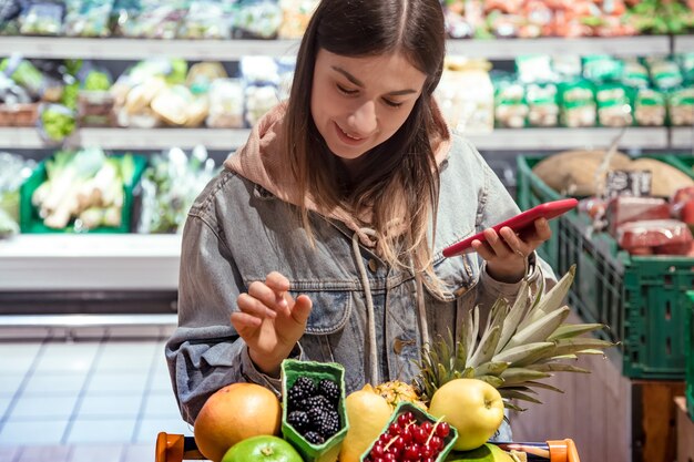 A young woman buys groceries in a supermarket with a phone in her hands.