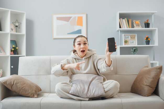 Young woman in casual clothing sitting on a couch at home interior showing smartphone pointing with index finger at it being amazed and surprised