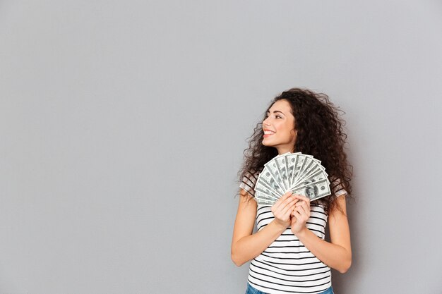 Young woman in casual holding fan of 100 dollar bills in hands looking aside with broad smile being glad over grey wall 