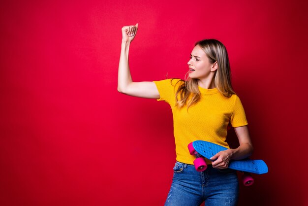 Young woman in casual outfit holding skateboard showing biceps while standing against red wall