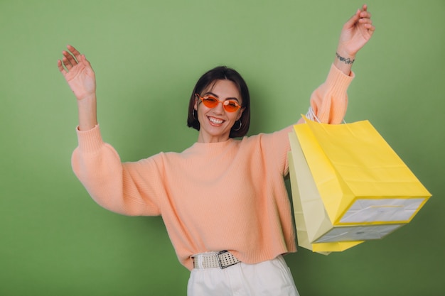 Free Photo young woman in casual peach sweater  isolated on green olive wall  holding shopping bags stylish in orange glasses jumping funny copy space