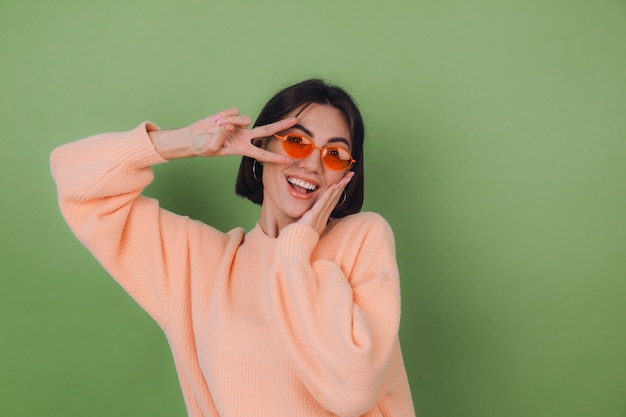 Young woman in casual peach sweater and orange glasses isolated on green olive wall   funny doing victory gesture copy space