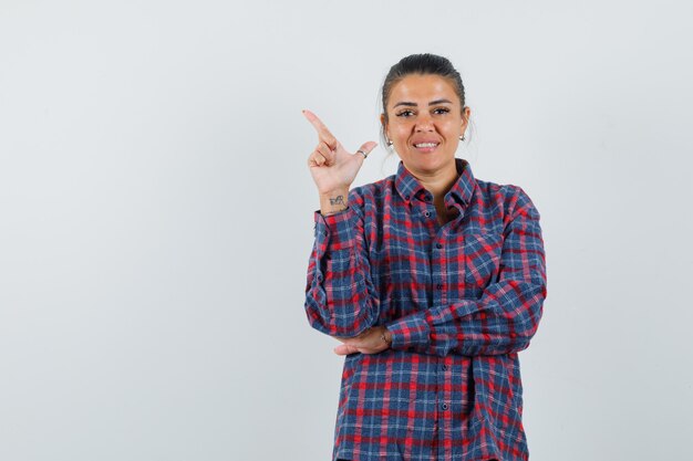 Young woman in checked shirt pointing upper left side with index finger and looking pretty , front view.