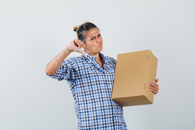 Free Photo young woman in checkered shirt holding box while showing thumb down and looking displeased.