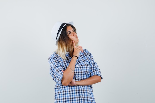 Young woman in checkered shirt looking away while pouting lips and looking pensive.