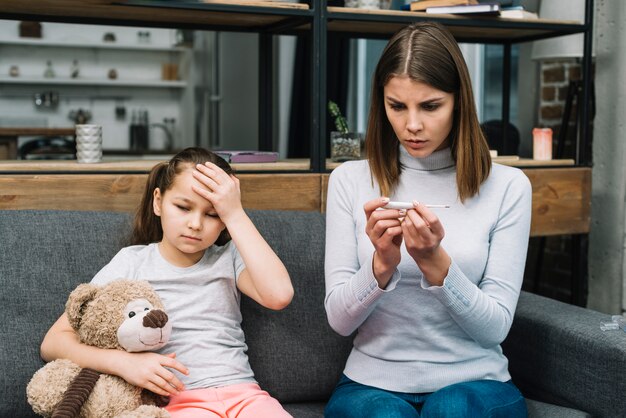 Young woman checking the temperature on thermometer sitting near the girl sitting with teddy bear