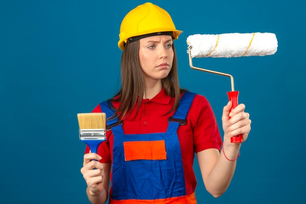 Free photo young  woman in construction uniform and yellow safety helmet holding brush and paint roller and looking at it with serious face standing on blue background