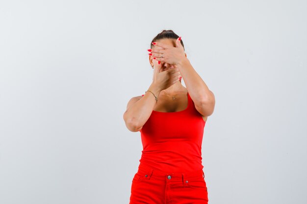 Young woman covering eyes and mouth with palms in red tank top, pants and looking upset , front view.