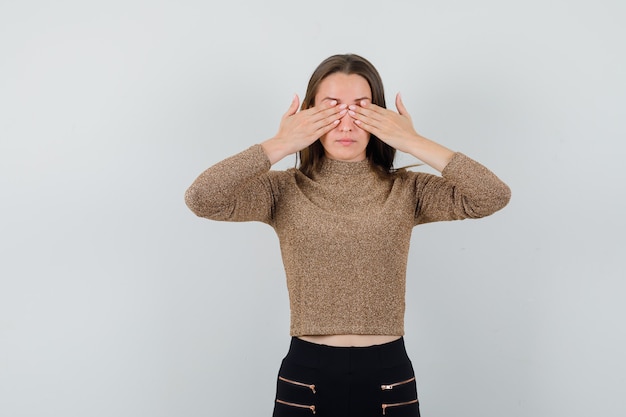 Free photo young woman covering eyes with hands in gold gilded sweater and black pants and looking serious , front view.