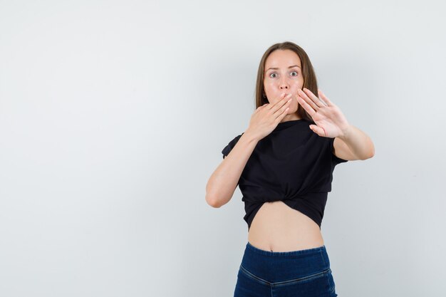 Young woman covering her mouth with hand while showing stop gesture in black blouse and looking troubled.