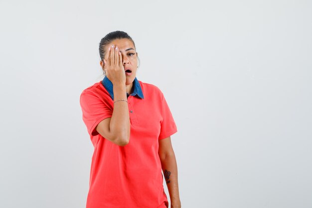 Young woman covering part of face with hand in red t-shirt and looking tired , front view.