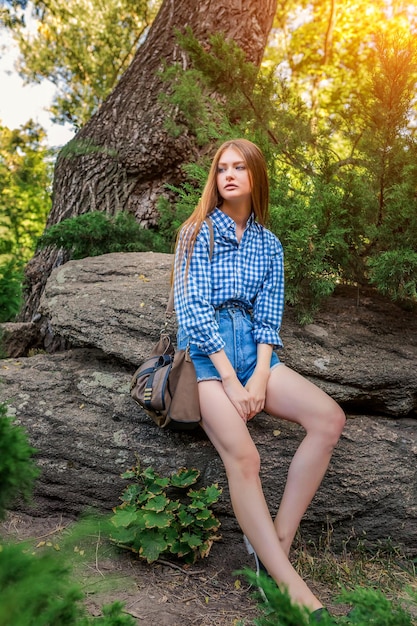 Young woman cute brunette girl sitting on stone, sun light flares of rays, looks into the distance on summer outdoor copy space background