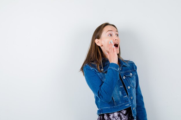 Young woman in denim jacket, dress shouting by keeping hand near mouth and looking excited , front view.