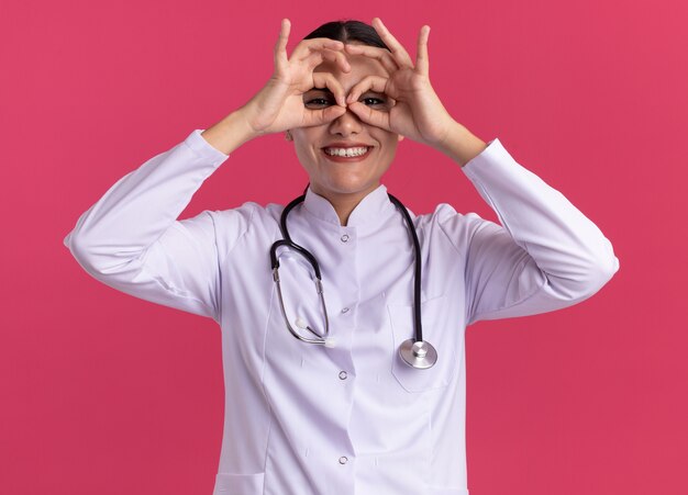 Young woman doctor in medical coat with stethoscope looking at front through fingers making binocular gesture smiling cheerfully standing over pink wall