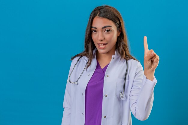 Young woman doctor in white coat with phonendoscope smiling confident and happy pointing finger up over isolated blue background