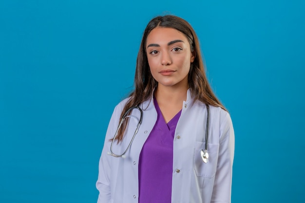 Young woman doctor in white coat with phonendoscope with serious face standing on blue isolated background