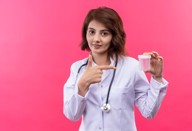 Young woman doctor in white coat with stethoscope holding test jar pointing finger to it smiling confident 