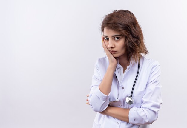 Young woman doctor in white coat with stethoscope looking at camera tired and bored leaning her head on arm 