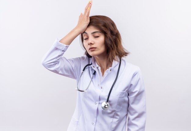 Young woman doctor in white coat with stethoscope looking tired and overworked with hand on head with closed eyes 