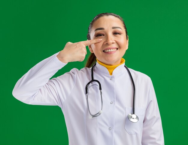 Young woman doctor in white medicine coat with stethoscope around neck pointing with index finger at her eye smiling cheerfully standing over green wall