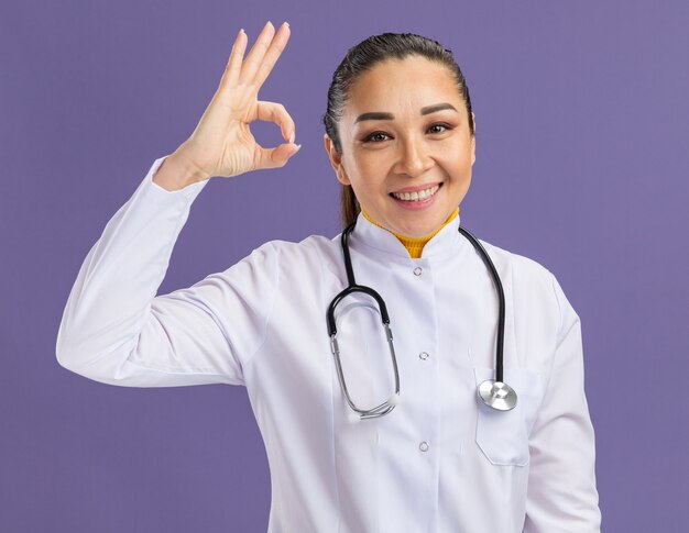 Young woman doctor in white medicine coat with stethoscope around neck  with smile on face doing ok sign standing over purple wall