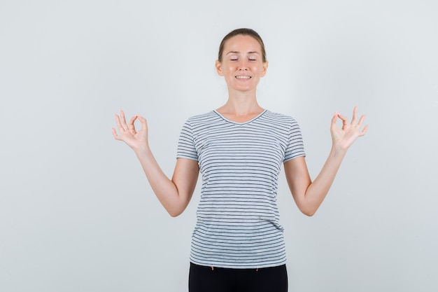 Young woman doing meditation with closed eyes in t-shirt, pants and looking relaxed. front view.