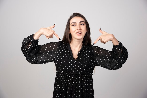 A young woman in dress pointing with two hands at herself.