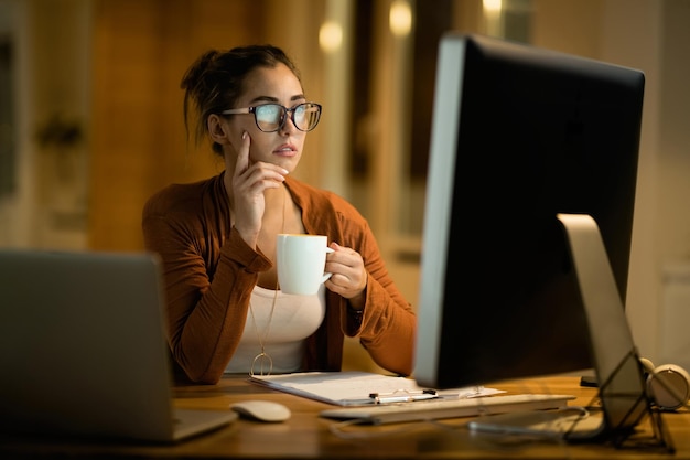 Young woman drinking coffee and thinking while working on desktop PC in the evening at home