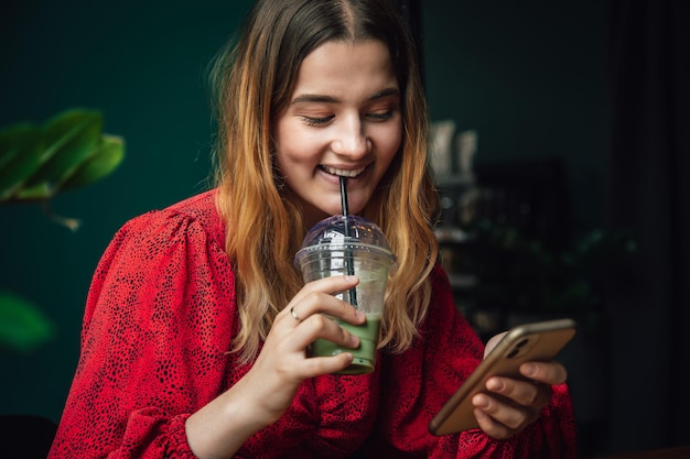 Young woman drinking green drink ice matcha latte in cafe and using smartphone