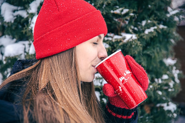 Free Photo a young woman drinks a hot drink from a red thermal cup in winter