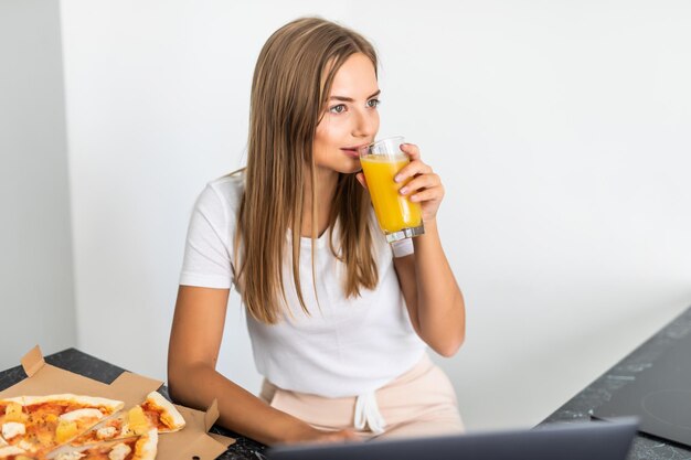 Young woman drinks juice and eating pizza and looking at laptop in the kitchen