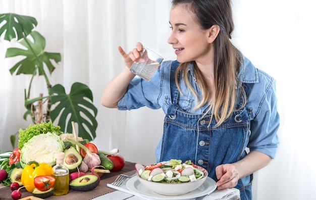 Free Photo a young woman drinks water at a table with vegetables on a light background , dressed in denim clothes. healthy food and drink concept .