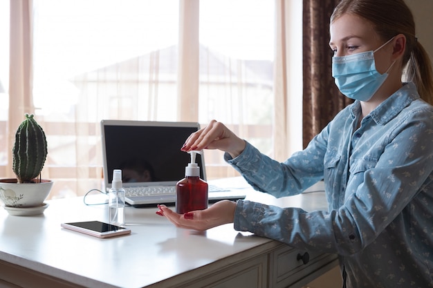 Young woman in face mask disinfecting gadgets surfaces on her workplace