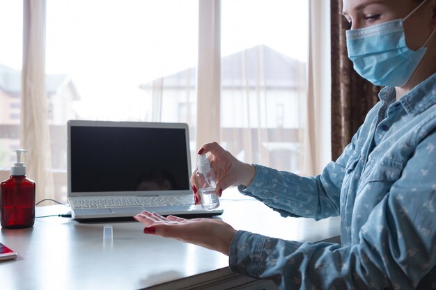 Young woman in face mask disinfecting gadgets surfaces on her workplace