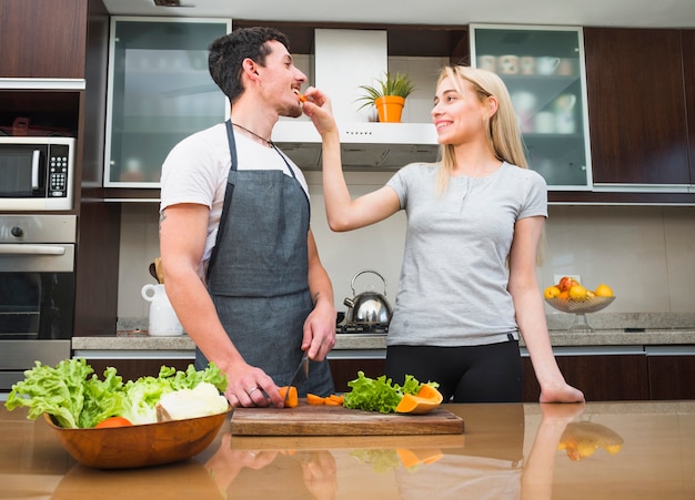 Young woman feeding carrot slice to her husband in the kitchen