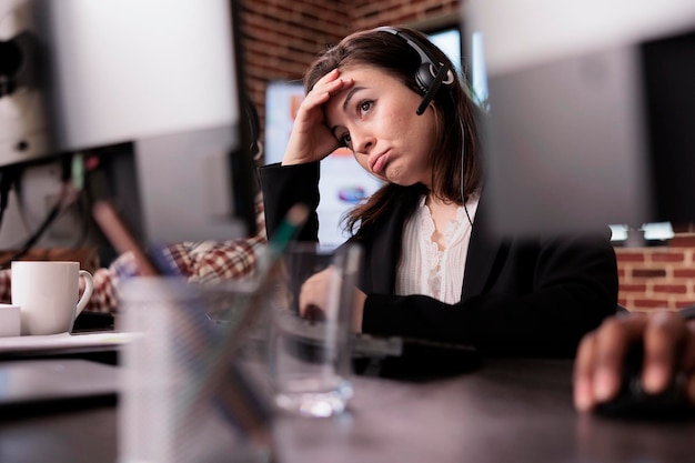 Young woman feeling tired at customer care support job, working on telemarketing call center at helpdesk. Female receptionist with helpline service helping clients on remote communication.