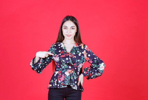 Young woman in floral shirt standing on red wall and presenting herself