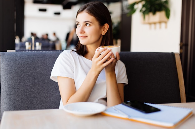 Young woman in gray dress sitting at table in cafe and writing in notebook. Student learning online.