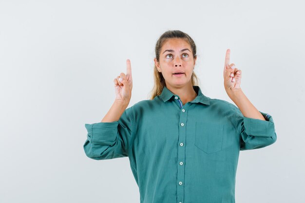 Young woman in green blouse pointing up with index fingers and looking pensive