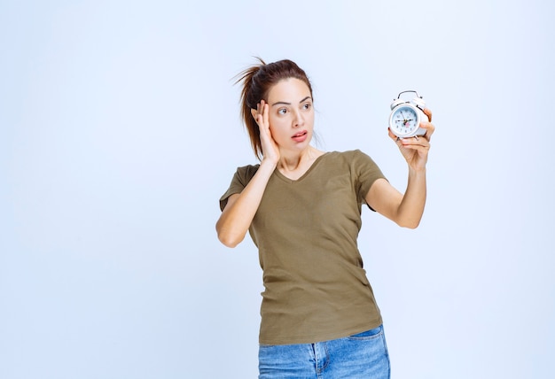 Young woman in green shirt holding an alarm clock and looks confused