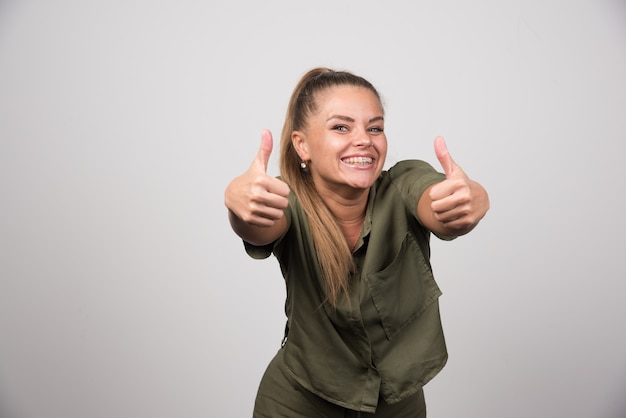 Young woman in green sweater giving thumbs up on gray wall.