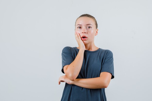 Young woman in grey t-shirt holding hand on cheek and looking surprised