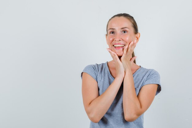 Young woman in grey t-shirt touching chin and looking cheerful , front view.