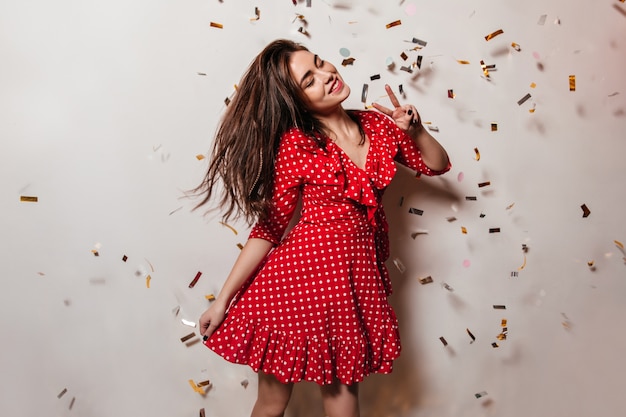 Young woman in high spirits is wearing red polka-dot dress. Girl with smile poses on wall of confetti and shows peace sign.