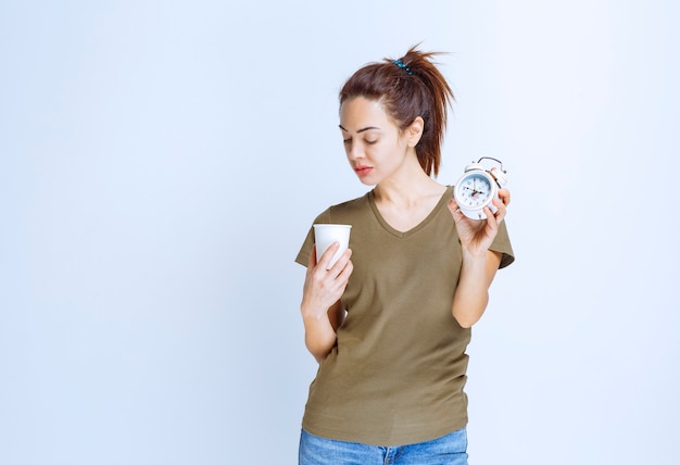 Young woman holding an alarm clock and a cup of drink pointing to the morning routine