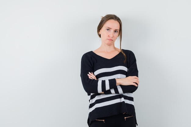 Young woman holding arms crossed in striped knitwear and black pants and looking serious