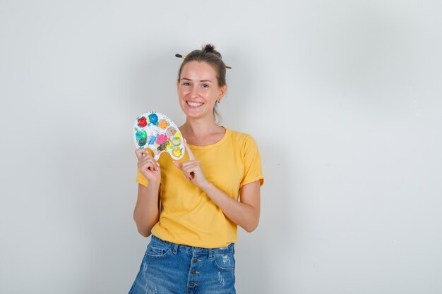 Young woman holding art palette in yellow t-shirt, jeans shorts and looking cheery